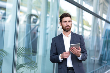 stylish bearded businessman in formal business suit standing working with tablet in hands on background modern office building outside. Man using smartphone or uses mobile phone outdoors city street