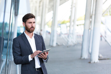 stylish bearded businessman in formal business suit standing working with tablet in hands on background modern office building outside. Man using smartphone or uses mobile phone outdoors city street