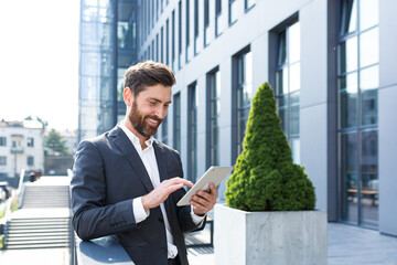 Cheerful and successful businessman works outdoors near the office holds a tablet, smiles reads...