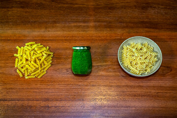 Stack of uncooked fusilli pasta, pesto, and cooked fusilli pasta dish. Wooden background.