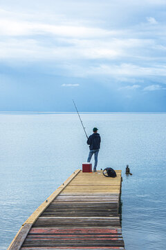 Young man fishing on dock with Caribbean Sea stretching to grey sky horizon.  Vertical format.  Salinas, Puerto Rico, USA.