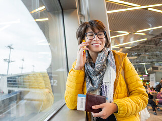 Smiling woman with passport and boarding pass is talking on her smartphone. Tourist is waiting for...
