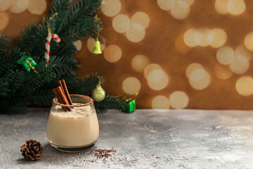 Christmas eggnog with cinnamon sticks on a background of fir branches decorated with toys, blurred bokeh. Glass of a festive winter drink on a concrete background with copy space.