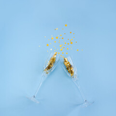 Two champagne glasses are decorated with gold star confetti on blue background. New year or birthday celebration concept.