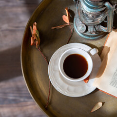 White cup of black coffee is on a metal table next to a book and a table lamp. Retro style. Square...