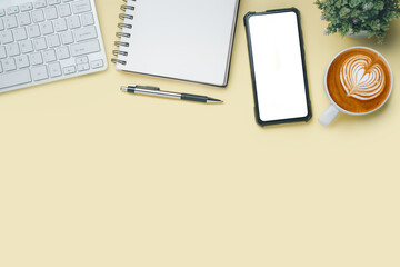 Office desk with computer, Blank screen smart phone, Pen, Notebook, Cup of coffee on yellow background, Top view with copy space, Mock up.