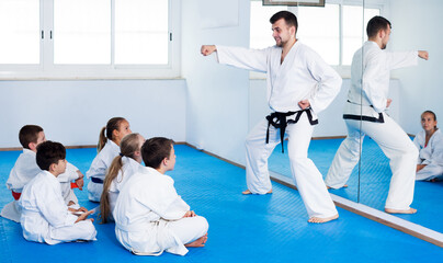 Fototapeta na wymiar Glad young male coach explaining new maneuvers to children in karate class