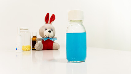 Medicinal syrup stands in the foreground behind it there are drops and vitamins and pills, as well as a stuffed plush toy bunny. Selective focus. Medicines for children. 