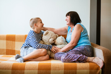 Kid plays with an old grandmother and teddy bear toy. Cheerful and smiling grandmother with her grandson. Cute toddler boy playing with senior woman sitting on sofa at home