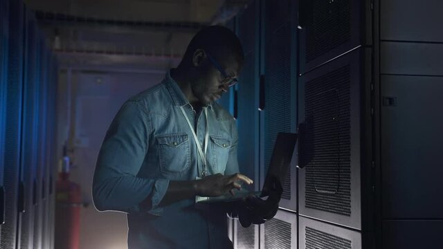 Young male engineer working on IT project with laptop in hands and standing in data office spbas. African American man checks equipment racks and holds computer, types and stands in dark interior