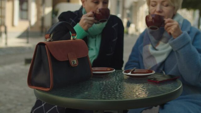 Focus on foreground, camera rotating around coffee shop table with handbag and cups of hot beverages against background of two mature adult women sitting at cafe table outdoors