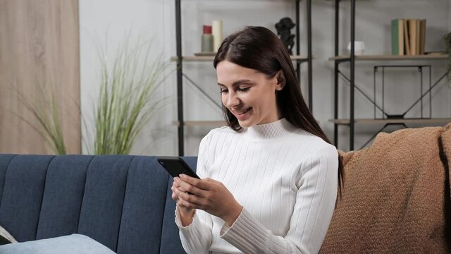 Pretty cheerful young brunette woman sitting on blue sofa uses phone smile at modern apartment texting share messages applications on social media enjoy free time feel happy mobile technology concept.