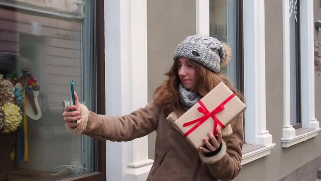 Girl in a brown coat making video selfie with gift box on the street near the showcase. Teenager using her camera on phone to make photo outside of the shop while shopping present for Christmas.