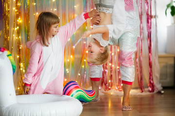 Funny mom in unicorn costume with toddler son and older sister together on multicolored rainbow background with lights with inflatable unicorn, children on holiday in style of unicorns, photo zone