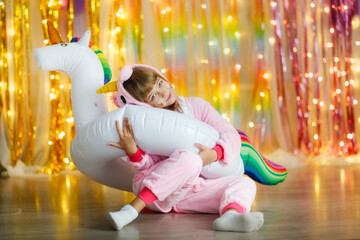 Fototapeta na wymiar Cute kid girl in unicorn costume on a multicolored rainbow background with lights with an inflatable unicorn, children at a party in the style of unicorns, photo zone