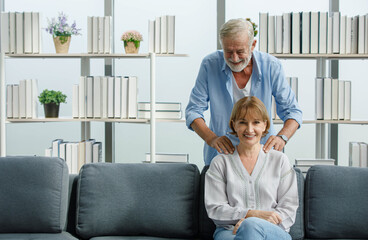 Caucasian old senior elderly gray bearded and hair male husband in casual wear standing behind massaging his lovely wife shoulder sitting smiling relaxing resting on sofa in living room at home