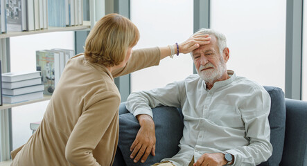 Caucasian old senior elderly supportive wife sitting use hand testing temperature from unhealthy gray bearded and hair husband forehead when he lying down on sofa get sick having cold and headache