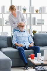 Caucasian old senior elderly lovely happy wife standing smiling behind do head massage for gray hair and bearded husband sitting close eyes resting relaxing comfortable on sofa in living room at home