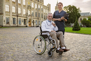 Fototapeta premium Full length shot of senior handicapped male doctor in wheelchair wearing lab coat holding glasses while posing together with young nurse assisting him outdoors