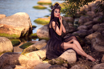 Dreaming Beautiful Brunette Female Ballerina In Black Dress Posing on Rocky Shore While Wearing Seashell Decorated Crown And Long Black Pearl Necklace.