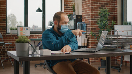 Employee looking at charts papers and laptop to work on business project and plan presentation. Man wearing face mask and working with data analysis on clipboard documents in office.