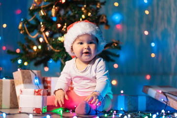 Winter seasonal holidays. Smiling toddler in a Christmas hat sitting with festive lights and xmas gifts. Indoor. The concept of Chrismas