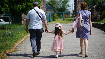 Fototapeta premium family walking in the park. mom dad and little girl walking holding hands. back view. daughter in a pink dress. friendly loving family concept, love. outdoor, walk in the fresh air, threesome