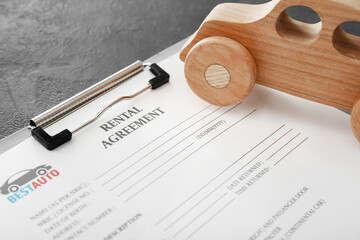 Clipboard with rental agreement and wooden car on dark background, closeup