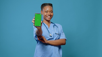 Smiling nurse showing green screen on mobile phone at camera. Medical assistant in uniform holding...