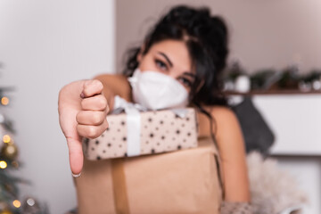 Upset sad woman opening wrapped ribbon gift box present,wear blue face disposable mask New Year evening, Christmas decorated tree. Hand gesture thumb down dislike lockdown holiday