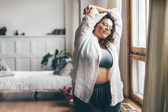 Curly haired overweight young woman in blue top and shorts with satisfaction on face accepts curvy body shape in stylish bedroom.