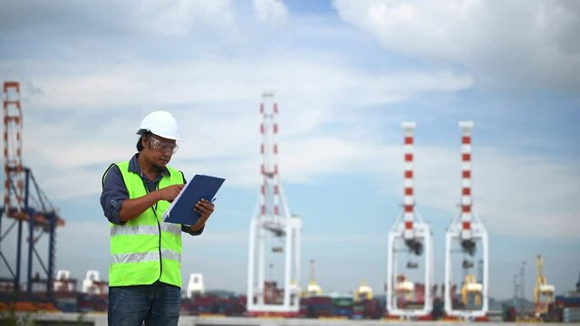 Engineer wearing safety uniform and helmet looking detail document on hand with logistic in habour backgroud.