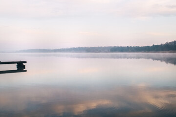 General plan of the pier for fishermen, which is located on a misty lake. The mist on the water creates the feeling of a magical effect. Clouds bounce off the surface. Place for text