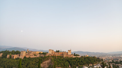 Aerial view of the famous Alhambra at sunset, Granada, Andalusia, Spain. White moon on the left. Pale blue sky on the background and view of the city on the right.