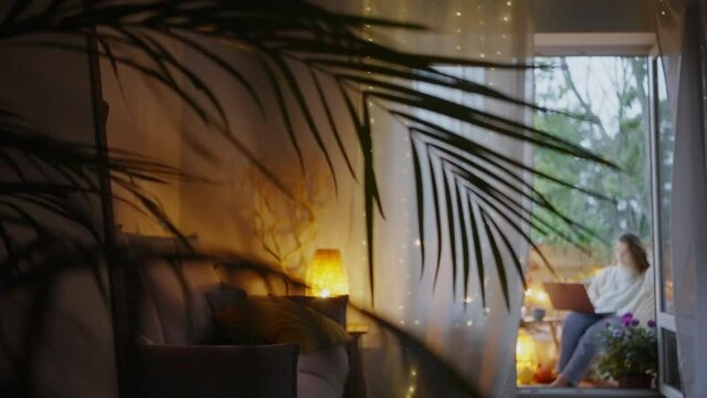 Curly haired young woman wearing warm clothes looks into laptop sitting on balcony decorated with lights and autumn attributes in evening view through palm branch
