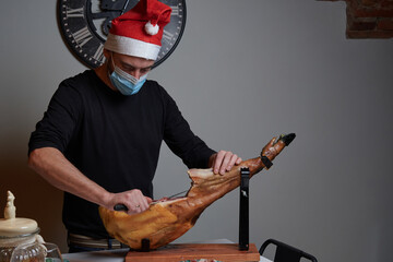 Professional ham cutter with protective face mask and Santa Claus hat cuts an Iberian ham with a...
