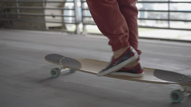 Young woman in red sports trousers makes tricks masterly with legs skateboarding with foot shoved under bridge against city in daytime backside view