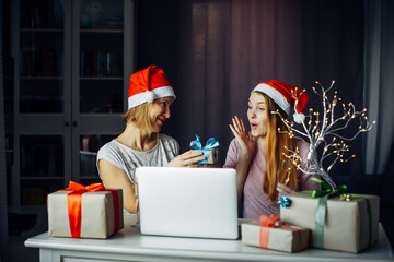 Fototapeta premium Two young women in Santa's hats exchange New Year's gifts in festive home interior. Joy of receiving a long-awaited gift for Christmas.