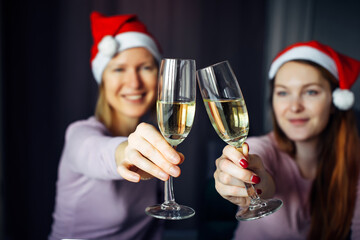 Christmas toast with glasses of champagne in hands. Blurred background: two girls in Santa Claus hats, clink glasses and wish Happy New Year. Love, friendship, family.