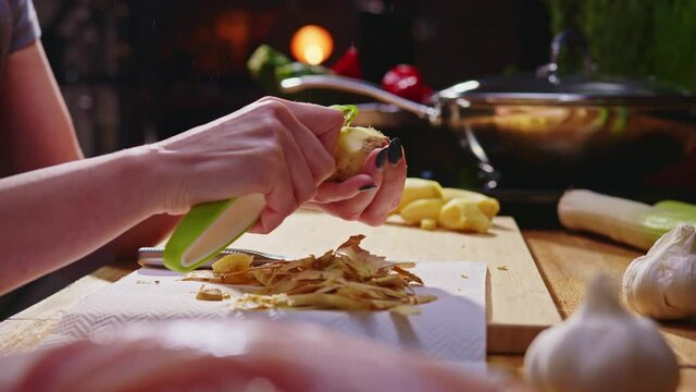Woman peeling ginger and vegetables for cooking on kitchen table. Closeup hands. Cosy dark room. Real, authentic cooking.