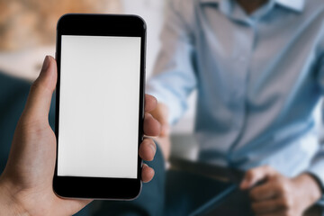 Hand holding white mobile phone with blank white screen in office.