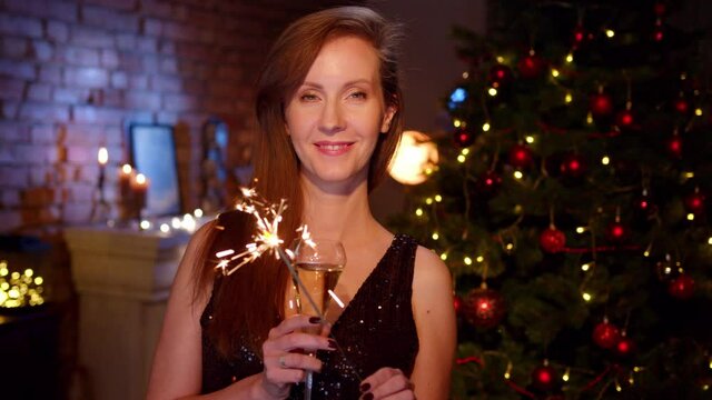 Happy woman wishing Merry Christmas and Happy New Year with glass of champagne and sparkler. Portrait of woman in elegant black dress at home with Christmas tree.