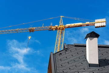 Construction site with orange tower crane and a closeup of a roof of a residential building with a chimney on a clear blue sky and clouds. Construction industry concept. Italy, Europe.