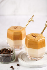Two glasses with trendy korean beverage dalgona coffee latte - whipped instant coffee, sugar and hot water and milk layers on a wooden board, white surface