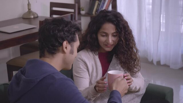 A Smiling Indian Husband offers a hot cup of coffee to a beautiful wife on a cold winter day. Happy man male and woman or female sitting on a couch enjoying a steamy cup of tea on a cozy morning.