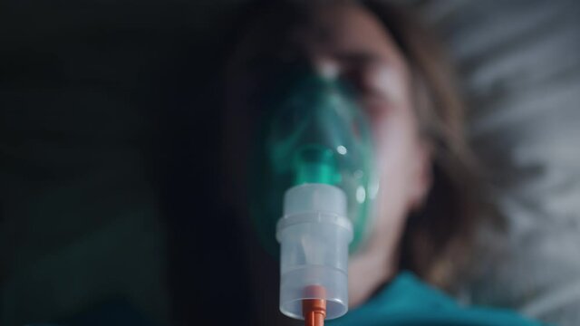 A female patient in medical mask is breathing oxygen support in a hospital next to heart rate machine. Unhealthy woman in coma. Getting mechanical ventilation. Concept of pandemic and therapy.