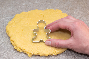The hand of the cook cutting out the shape of gingerbread men from raw rolled dough on a gray background.