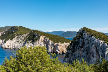 Fototapeta premium Greenery, high steep rocky cliffs, sunny sea shore on a bright clear blue day in Greece. Scenic travel destination. Lefkada island, vivid Ionian sea coast