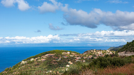 Fototapeta premium Picturesque traditional greek village with tiled roofs on green hills with scenic cloudscape near blue sea shore on Lefkada island, Greece. Travel Europe in summer