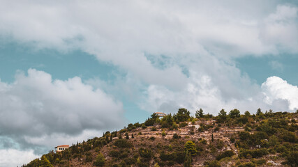 Scenic vibrant landscape with traditional Greek houses on green high hills on blue scenic sky with dramatic cloudscape on Lefkada island, Greece. Travel Europe in summer. Color graded
